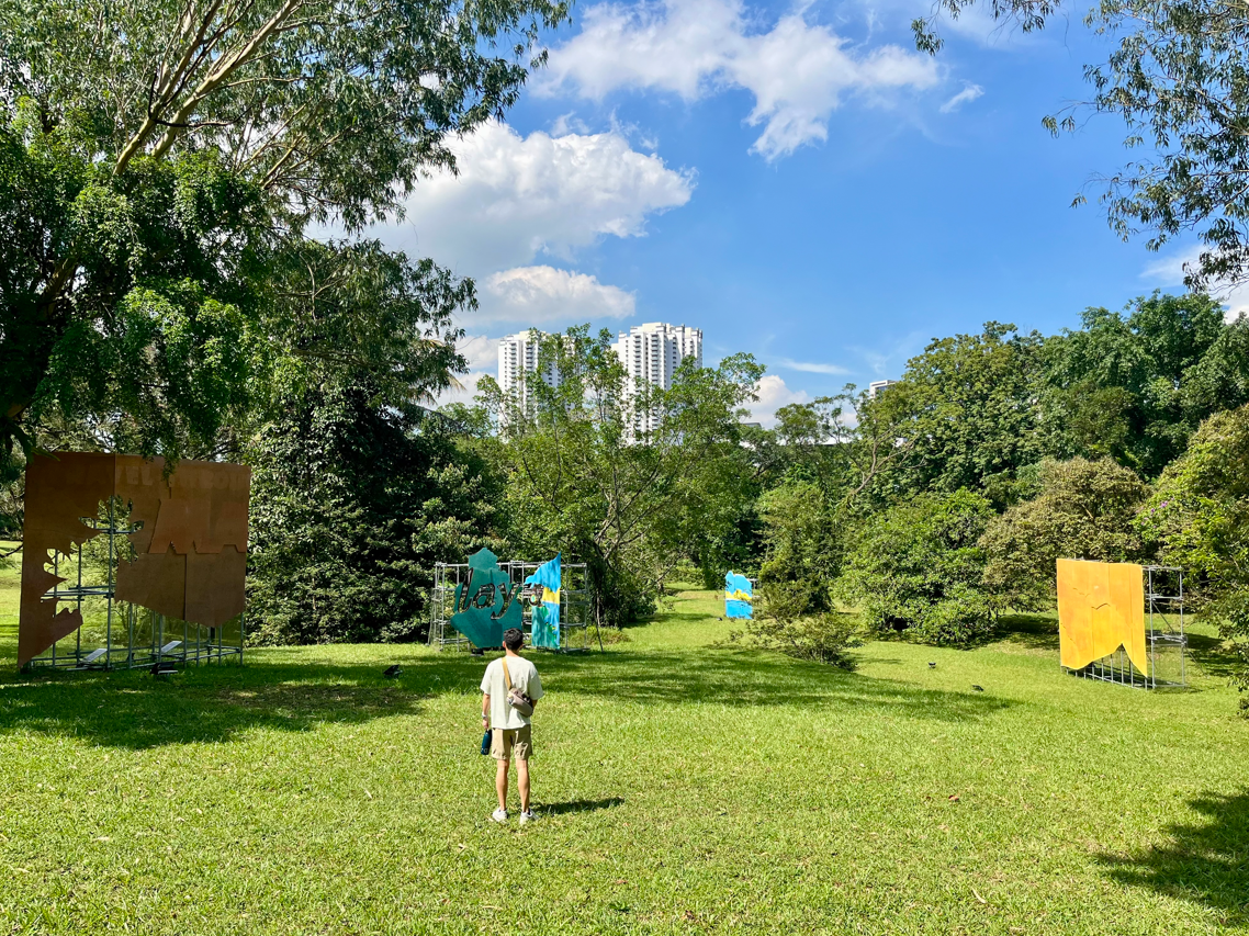 A person standing on green grass looking at outdoor art installations in a park on a sunny day. Buildings in the background.